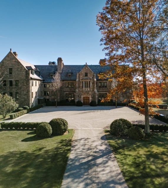 Stone campus-style building with a central arched doorway, symmetry, and a gravel courtyard edged by trimmed hedges under a clear blue sky.