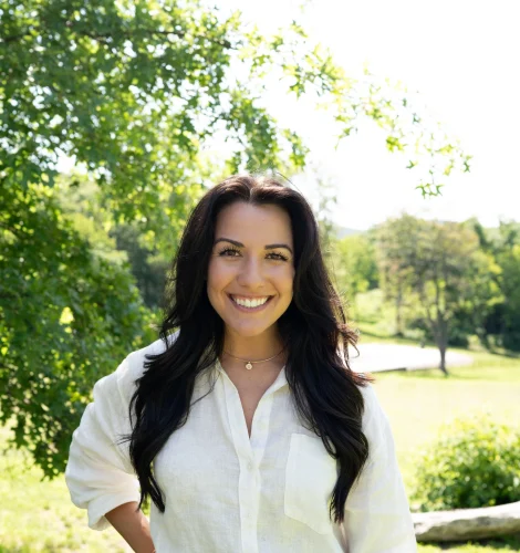 Smiling woman with long dark hair in a white blouse and jeans, standing in a sunlit park with green trees behind her.