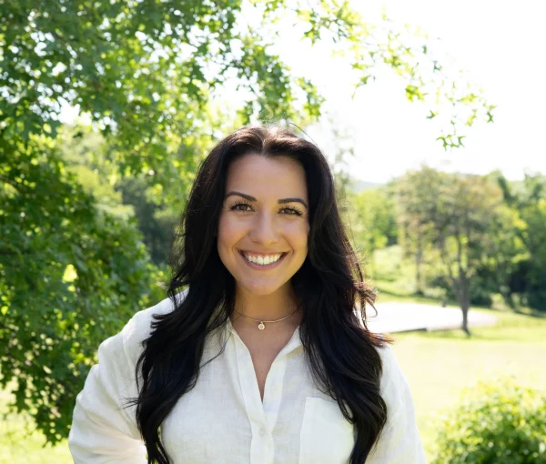 Smiling woman with long dark hair in a white blouse and jeans, standing in a sunlit park with green trees behind her.