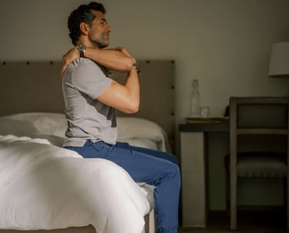 Man sits on edge of bed in hotel room, stretching his shoulders with arms crossed over chest.