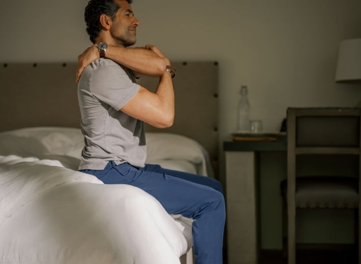 Man sits on edge of bed in hotel room, stretching his shoulders with arms crossed over chest.