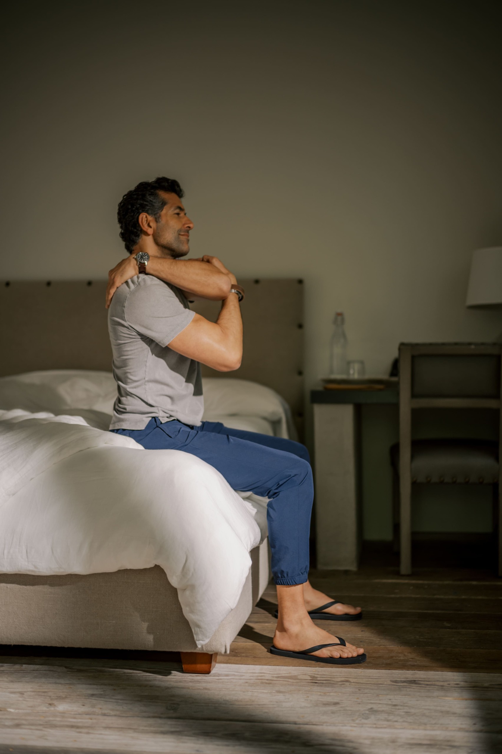 Man sits on edge of bed in hotel room, stretching his shoulders with arms crossed over chest.