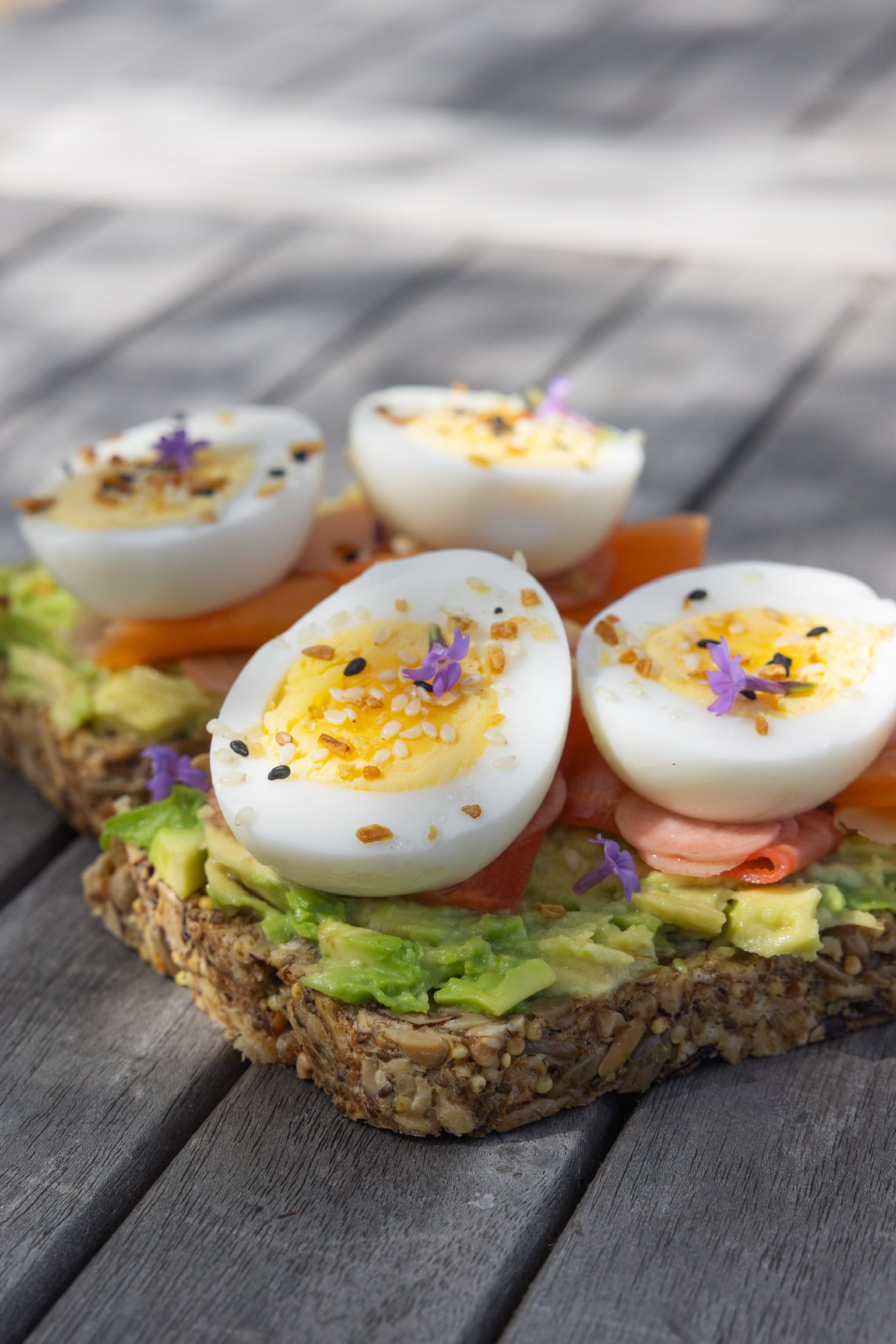Open-faced toast topped with mashed avocado, tomato slices, and halved deviled eggs sprinkled with sesame seeds and edible purple flowers on a wooden table.