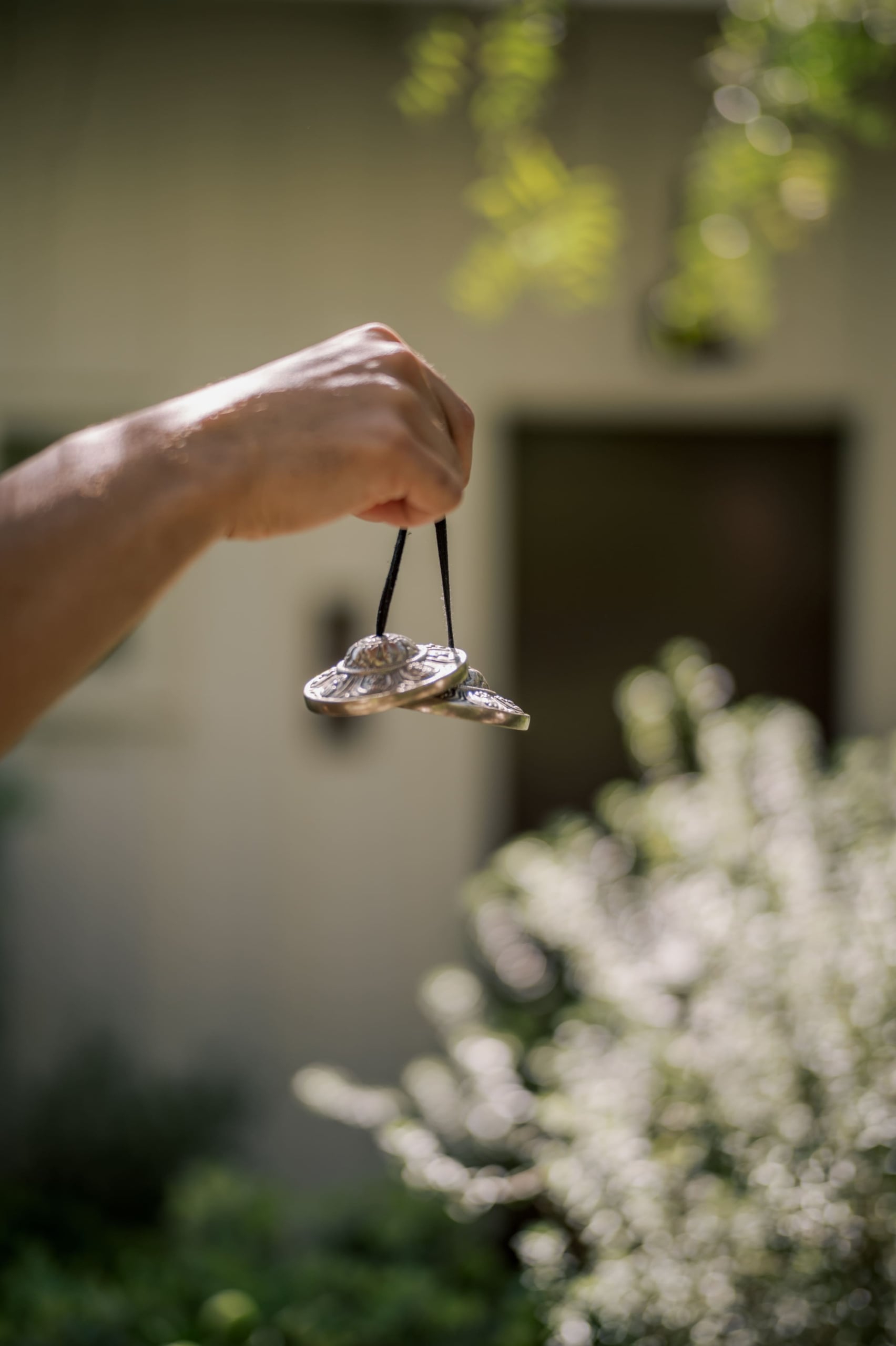 Close-up of a hand holding silver metal bells on a black cord, outdoors with green bokeh in the background.