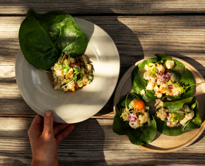 Hand holding a beige plate with a small chopped salad resting on a large green leaf on a wooden table.