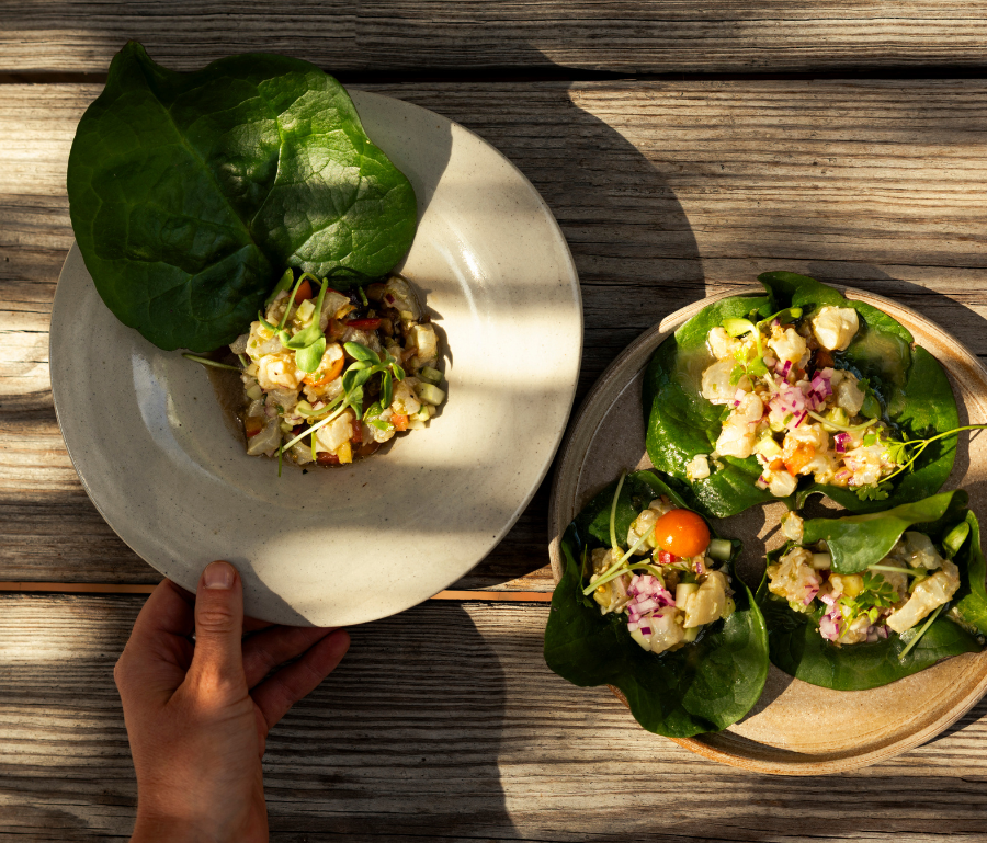 Hand holding a beige plate with a small chopped salad resting on a large green leaf on a wooden table.
