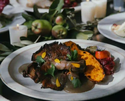 Close-up of a plated gourmet dish with roasted mushrooms, colorful vegetables, and fresh herbs on a white plate, candlelit table in background.