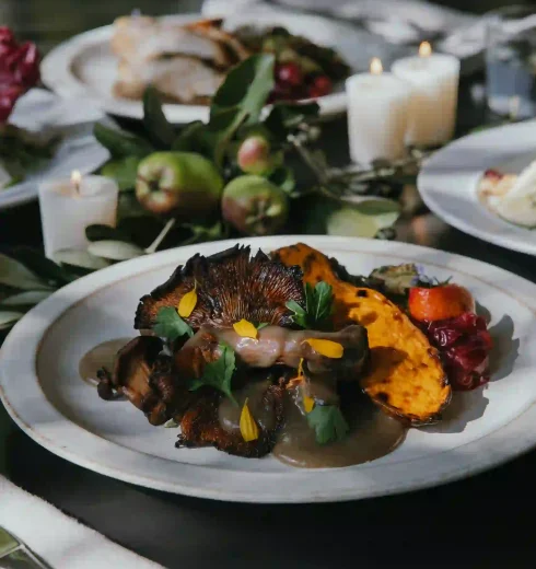 Close-up of a plated gourmet dish with roasted mushrooms, colorful vegetables, and fresh herbs on a white plate, candlelit table in background.
