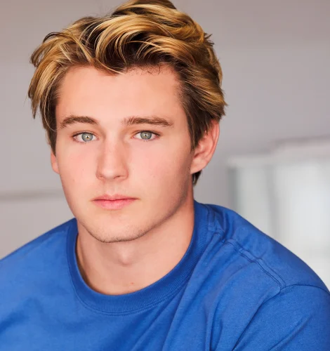 Close-up portrait of a young man with blond hair and blue eyes wearing a blue T-shirt against a neutral background.