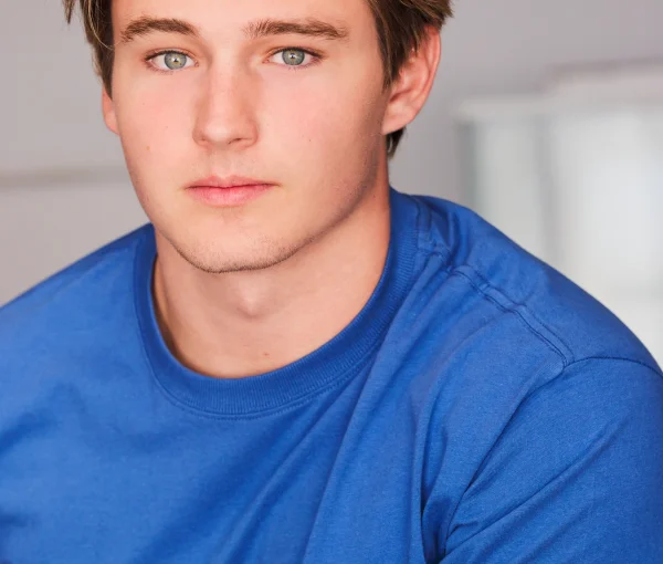 Close-up portrait of a young man with blond hair and blue eyes wearing a blue T-shirt against a neutral background.