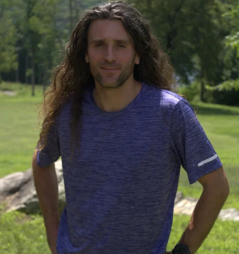 Man with long brown hair standing in a sunny park, wearing a blue heathered shirt and hands on hips.