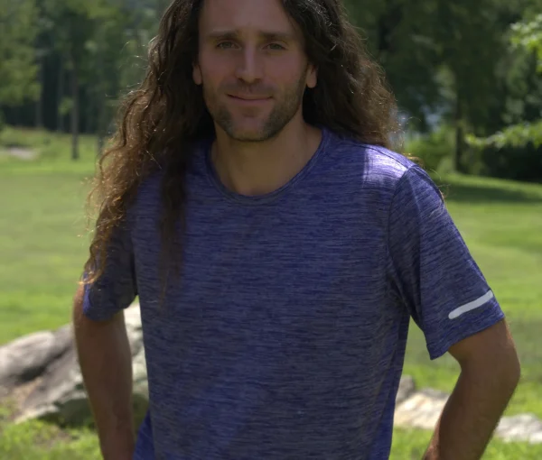 Man with long brown hair standing in a sunny park, wearing a blue heathered shirt and hands on hips.