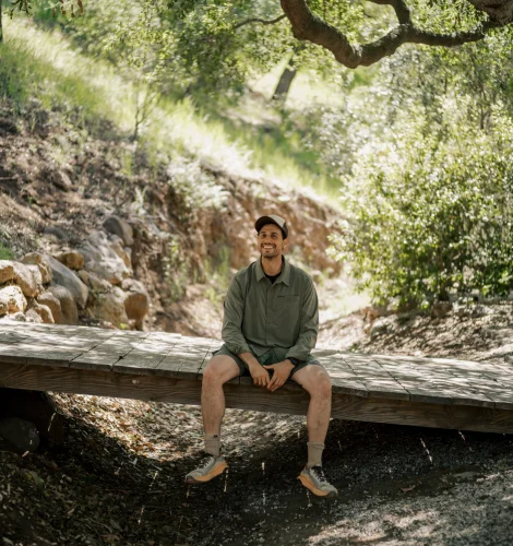 Man sits on a wooden bridge in a sunlit forest, smiling, wearing a green shirt and cap with trees overhead.