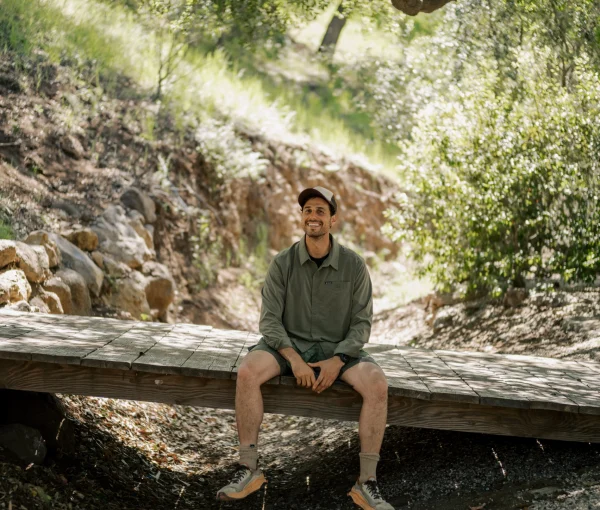 Man sits on a wooden bridge in a sunlit forest, smiling, wearing a green shirt and cap with trees overhead.