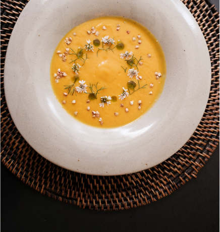Creamy orange soup in a white bowl, garnished with small white flowers, dill-like herbs, and sprinkled seeds on a woven placemat.