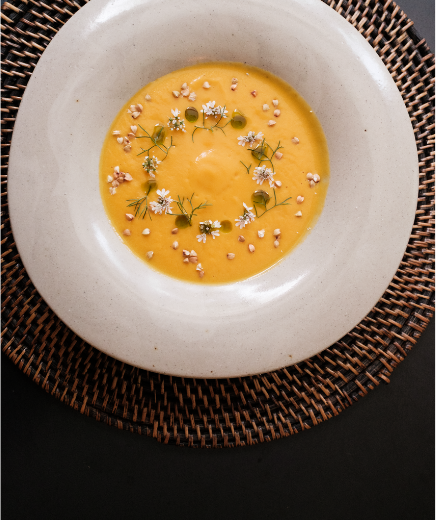 Creamy orange soup in a white bowl, garnished with small white flowers, dill-like herbs, and sprinkled seeds on a woven placemat.