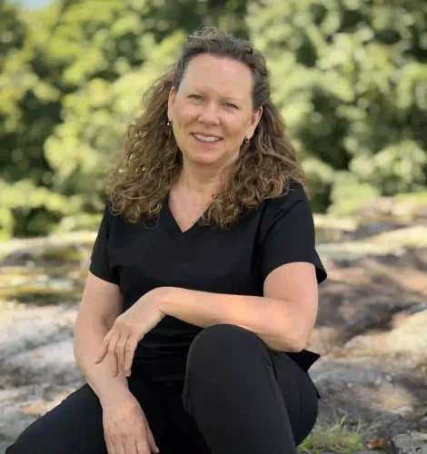 Woman sitting on stone steps outdoors, smiling, wearing a black top and black pants with green foliage in the background.