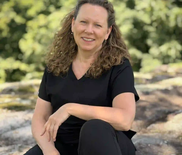 Woman sitting on stone steps outdoors, smiling, wearing a black top and black pants with green foliage in the background.