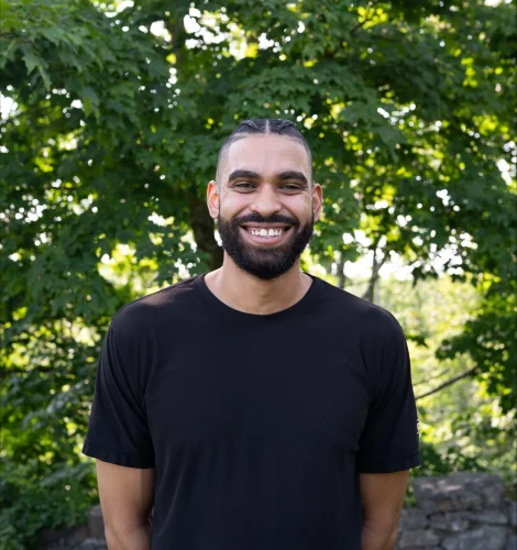 Smiling man with a beard in a black t-shirt standing outdoors with green trees and a stone wall behind him.