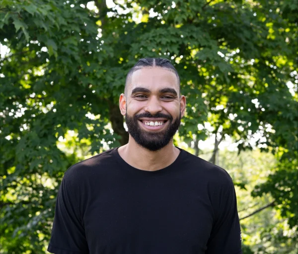 Smiling man with a beard in a black t-shirt standing outdoors with green trees and a stone wall behind him.