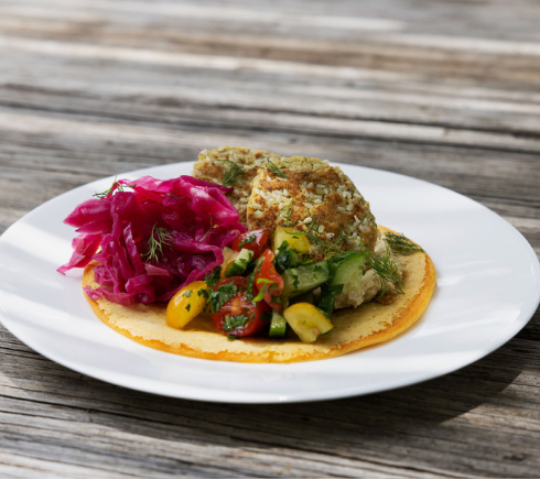 Crispy taco on a corn tortilla topped with diced vegetables and herbs, with pink pickled cabbage slaw on the side, served on a white plate.