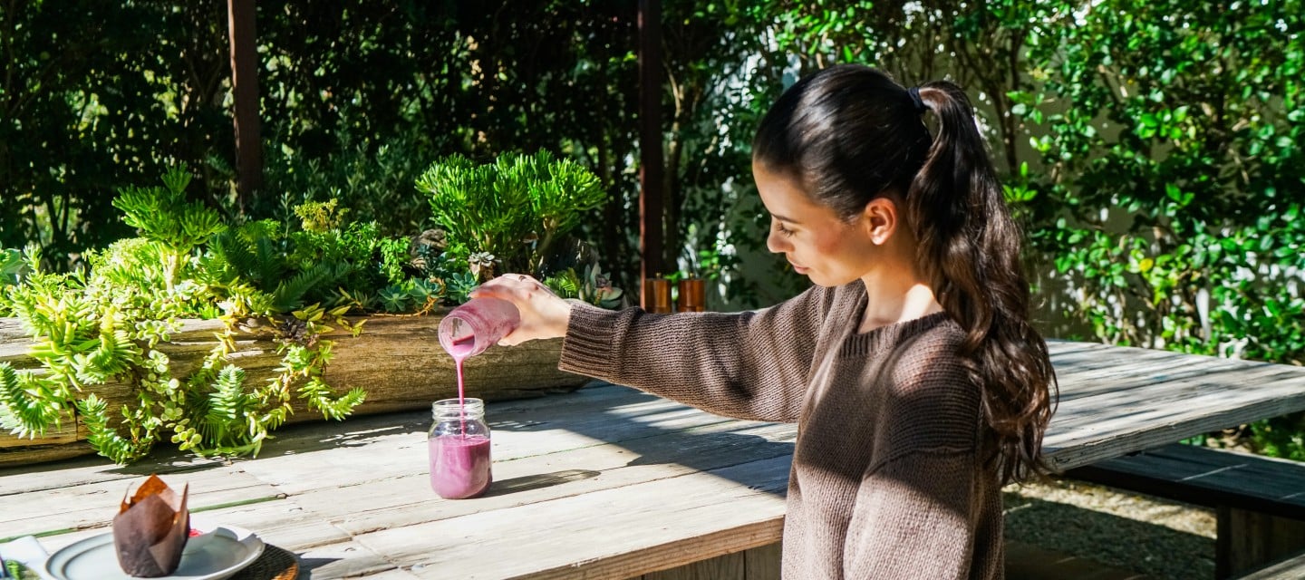 Woman sits at a wooden outdoor table, pouring a pink smoothie from a bottle into a jar amid potted succulents and greenery.