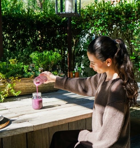 Woman sits at a wooden outdoor table, pouring a pink smoothie from a bottle into a jar amid potted succulents and greenery.
