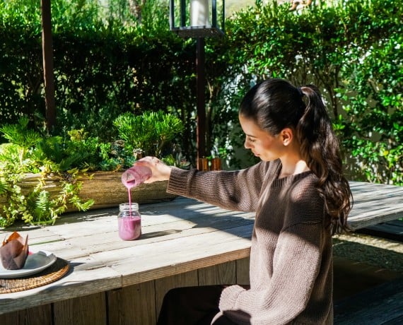 Woman sits at a wooden outdoor table, pouring a pink smoothie from a bottle into a jar amid potted succulents and greenery.