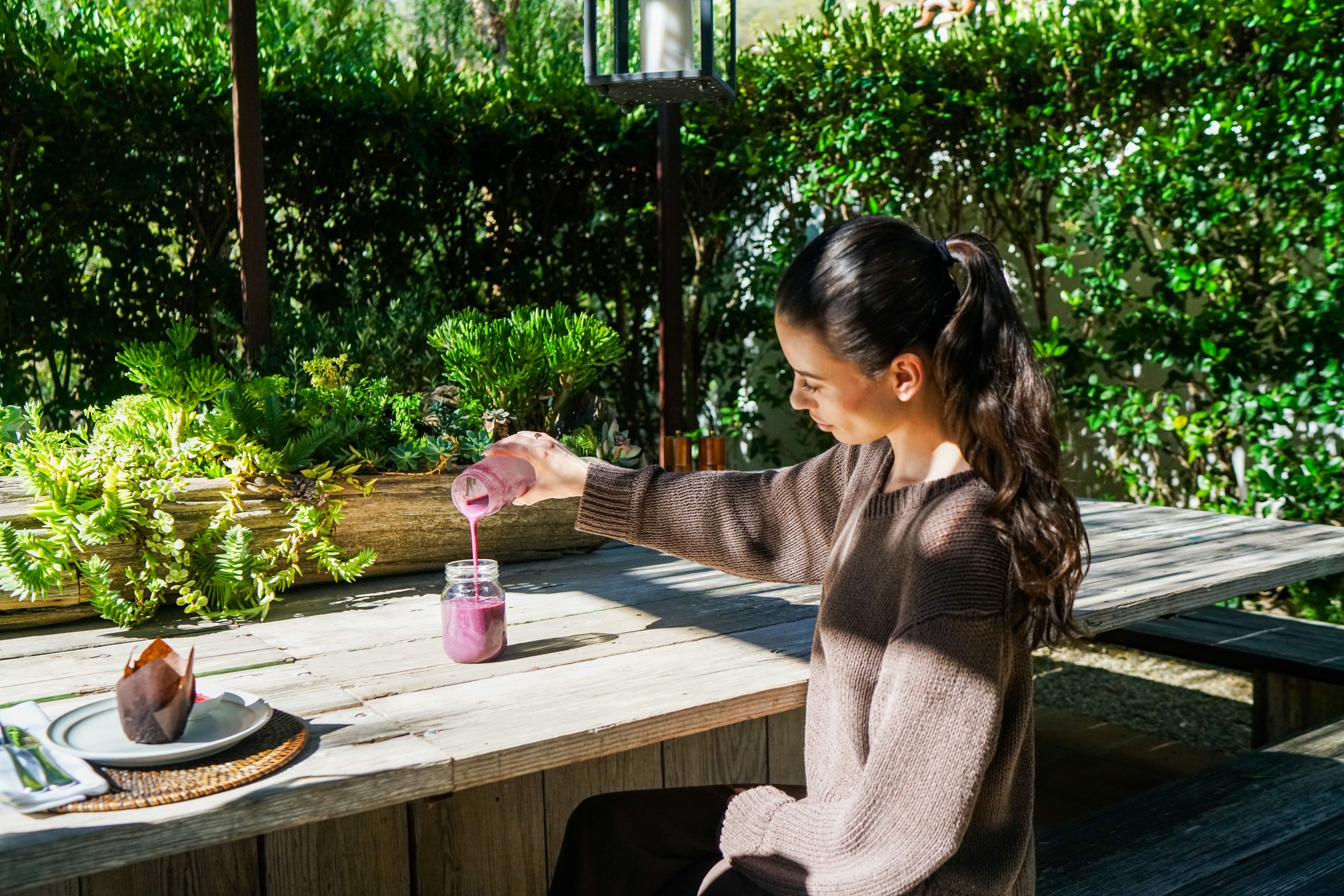 Woman sits at a wooden outdoor table, pouring a pink smoothie from a bottle into a jar amid potted succulents and greenery.