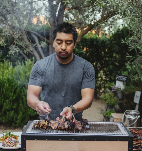 Man wearing a gray shirt tending meat skewers on an outdoor grill in a backyard garden, with trees in the background.