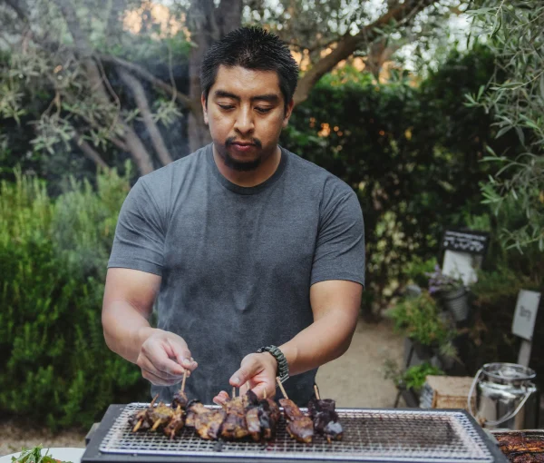 Man wearing a gray shirt tending meat skewers on an outdoor grill in a backyard garden, with trees in the background.