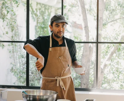 Man wearing an apron and cap stands at a kitchen counter with bowls and ingredients, preparing food by a large window.