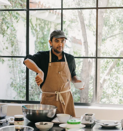 Man wearing an apron and cap stands at a kitchen counter with bowls and ingredients, preparing food by a large window.