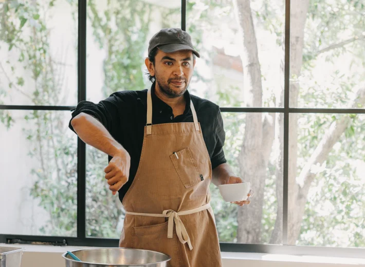 Man wearing an apron and cap stands at a kitchen counter with bowls and ingredients, preparing food by a large window.