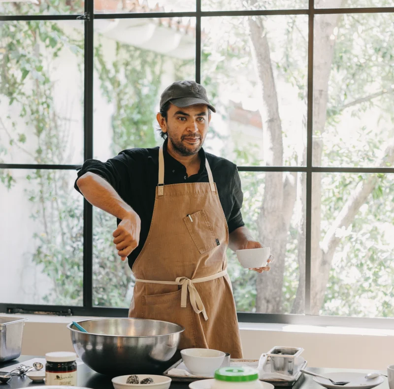 Man wearing an apron and cap stands at a kitchen counter with bowls and ingredients, preparing food by a large window.