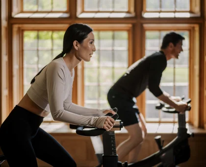 Two people ride stationary bikes in a sunlit wood-paneled room during a workout class.