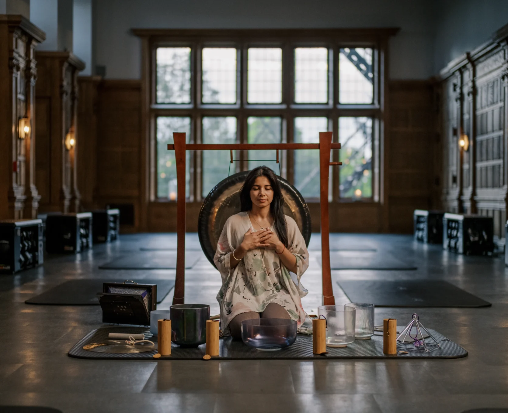 Woman meditates with hands on chest in front of a large gong, inside a dim, wood-paneled hall.