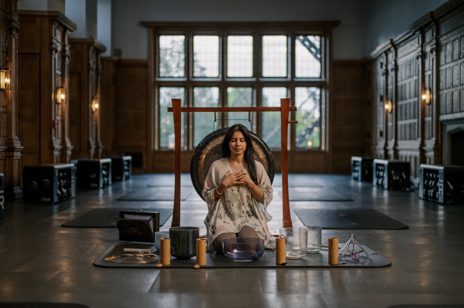 Woman meditates with hands on chest in front of a large gong, inside a dim, wood-paneled hall.