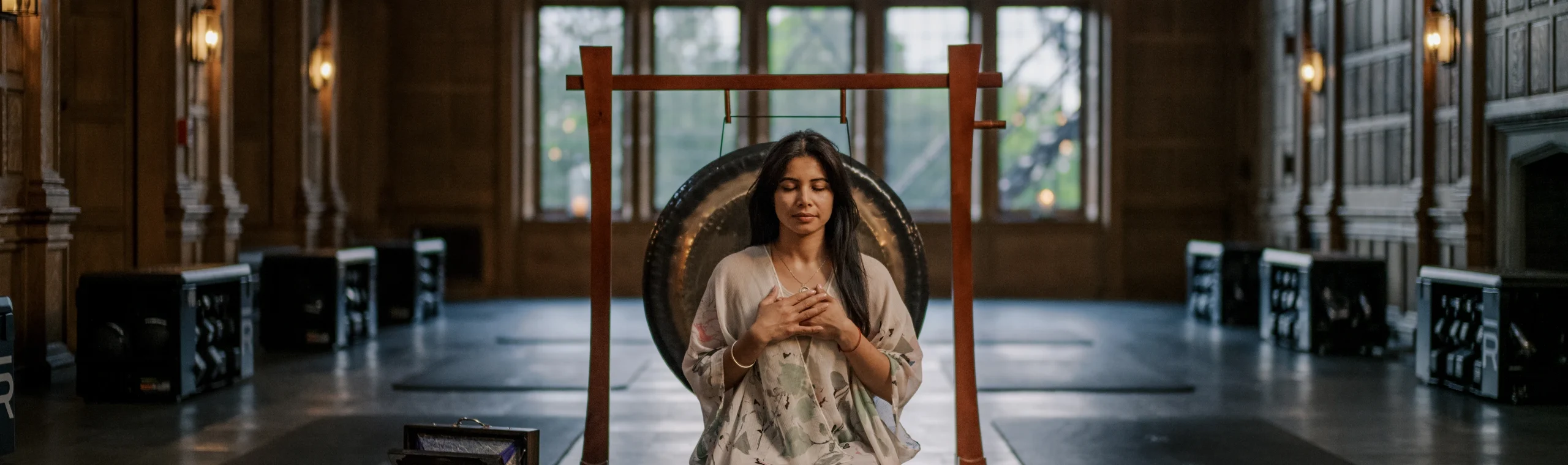 Woman meditates with hands on chest in front of a large gong, inside a dim, wood-paneled hall.