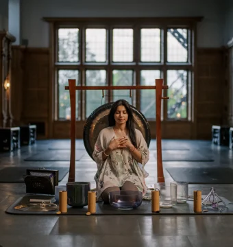 Woman meditates with hands on chest in front of a large gong, inside a dim, wood-paneled hall.