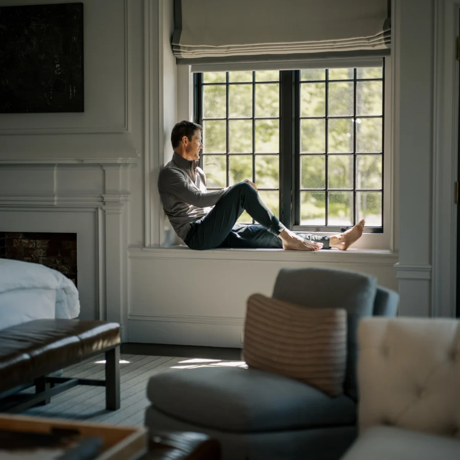 Man sits cross-legged on a wide window sill in a bright living room, gazing outside at greenery.