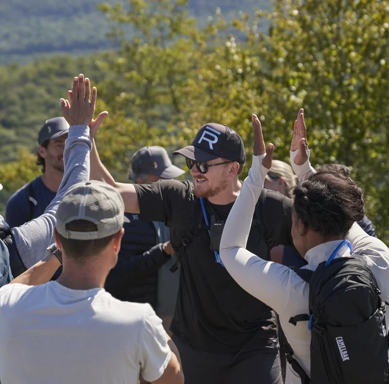 Group of hikers outdoors giving high-fives in a mountainous area with trees in the background and blue sky.