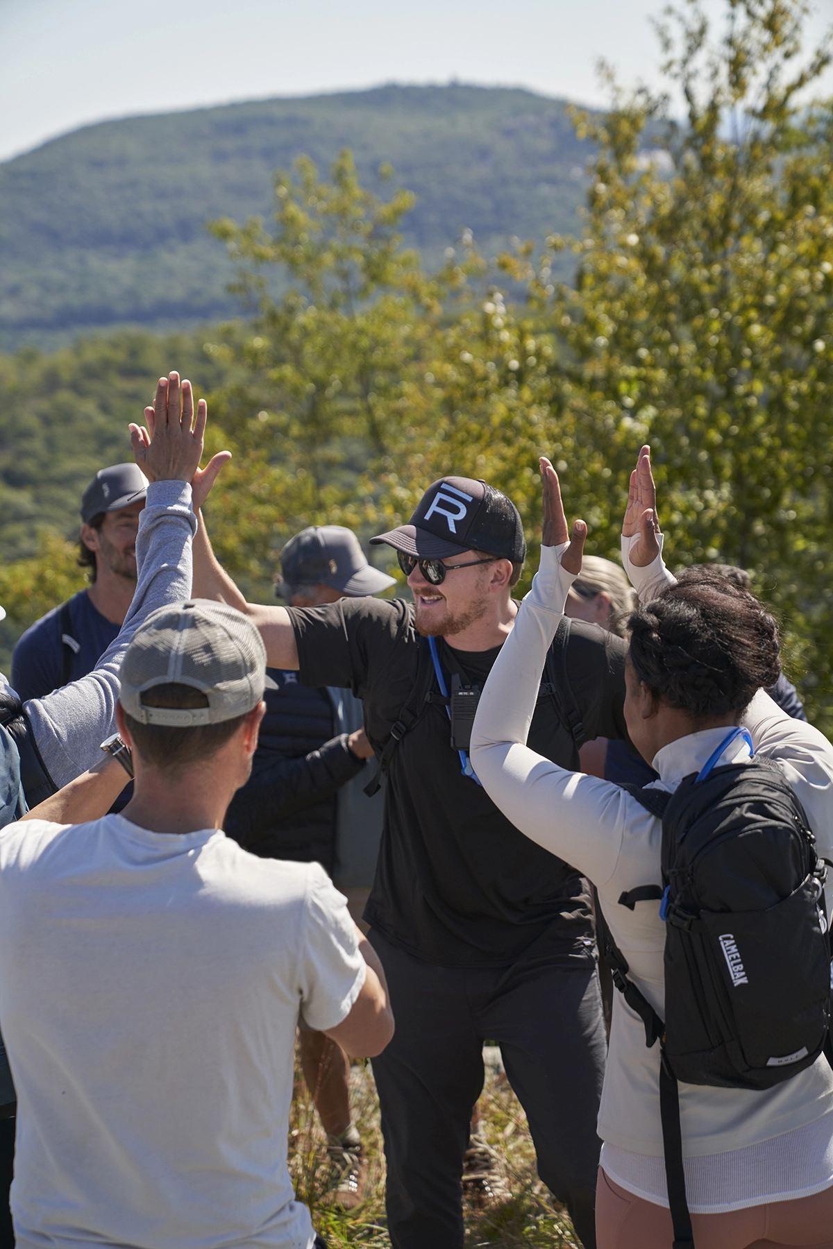 Group of hikers outdoors giving high-fives in a mountainous area with trees in the background and blue sky.