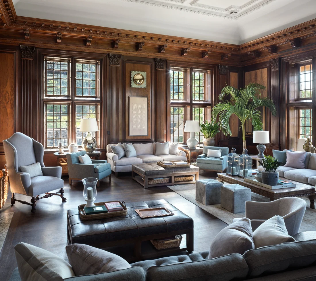 Luxurious lounge with dark wood paneling, large grid windows, and blue-gray sofas around a leather-tufted ottoman.