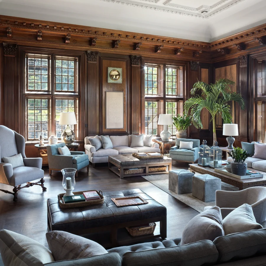 Luxurious lounge with dark wood paneling, large grid windows, and blue-gray sofas around a leather-tufted ottoman.