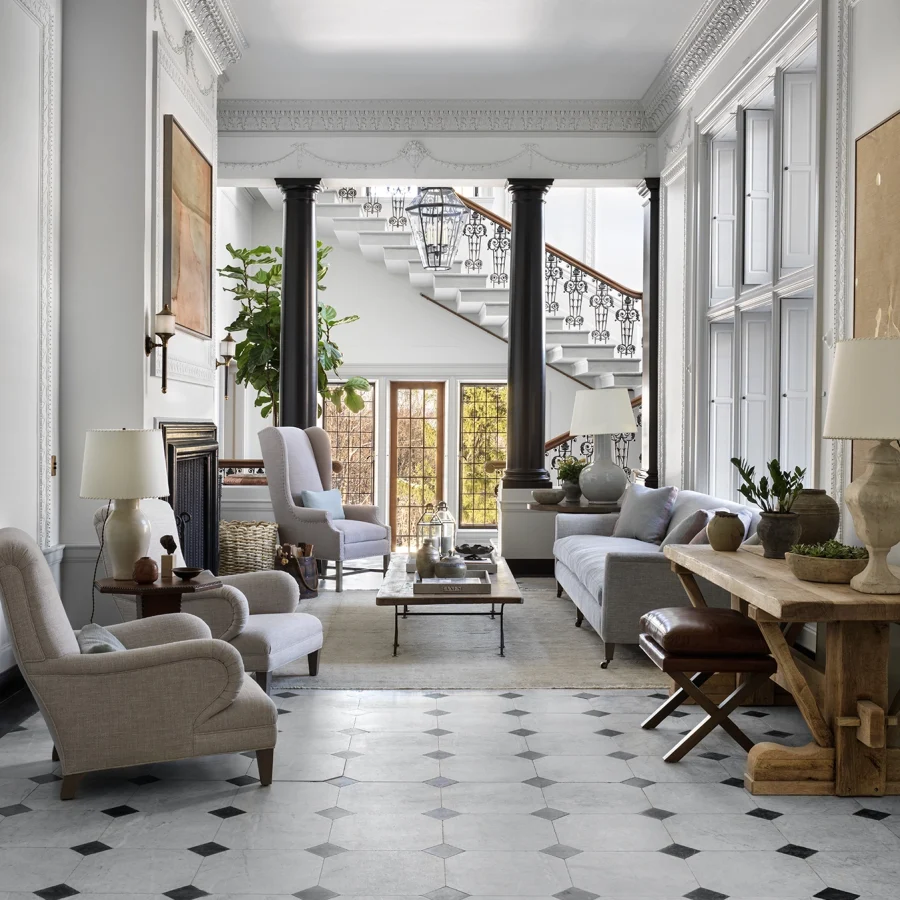 Luxurious living room with white walls, ornate molding, black columns, and a grand staircase visible in the background.
