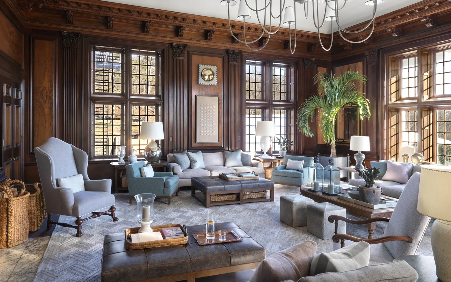 Luxurious living room with dark wood paneling, oversized windows, and a grand chandelier overhead.