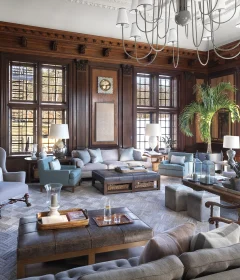 Luxurious living room with dark wood paneling, oversized windows, and a grand chandelier overhead.