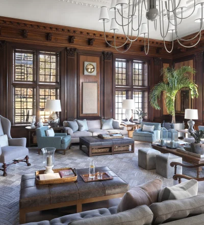 Luxurious living room with dark wood paneling, oversized windows, and a grand chandelier overhead.