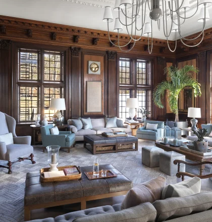 Luxurious living room with dark wood paneling, oversized windows, and a grand chandelier overhead.
